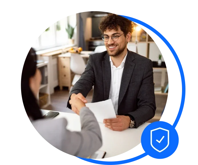 Male recruiter wearing a white shirt with black jacket, glasses shaking a females hand. He is holding a piece of paper in the other hand and they are sat down at a desk.